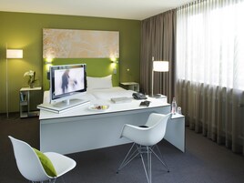 Habitación de hotel con cama, escritorio con computadora, silla, televisor y ventana con cortinas.