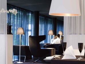 A modern hotel room with a dark blue curtain backdrop, a black armchair, a round table with a white napkin, a wine glass, and a lamp.