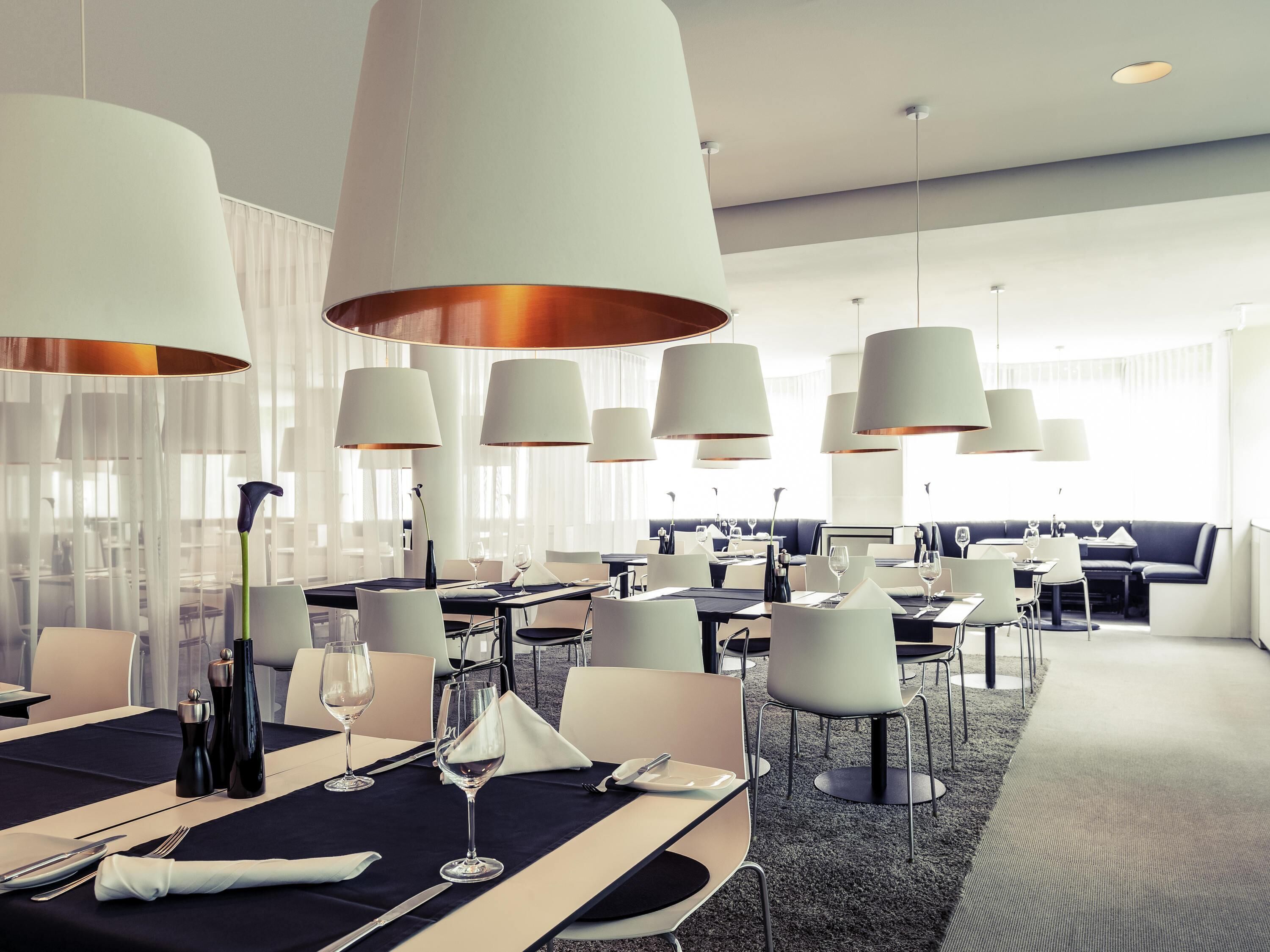 Un espace de restauration moderne dans un hôtel, avec des tables dressées pour le repas, de grandes lampes suspendues et un coin salon avec un canapé.