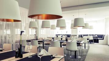 Un espace de restauration moderne dans un hôtel, avec des tables dressées pour le repas, de grandes lampes suspendues et un coin salon avec un canapé.