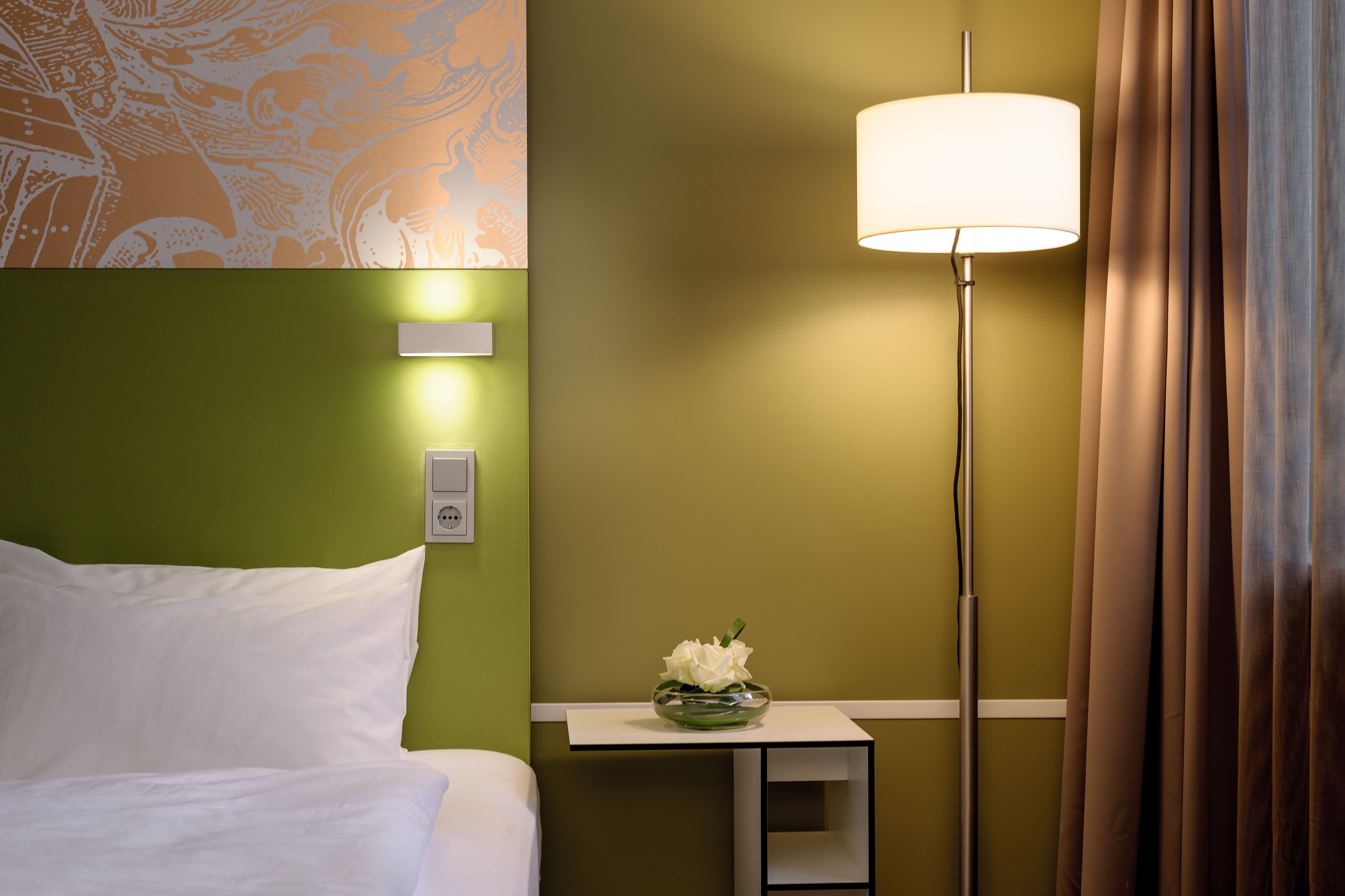 Habitación de hotel con cama, mesita de noche con lámpara, un jarrón con flores y un diseño decorativo en la pared.