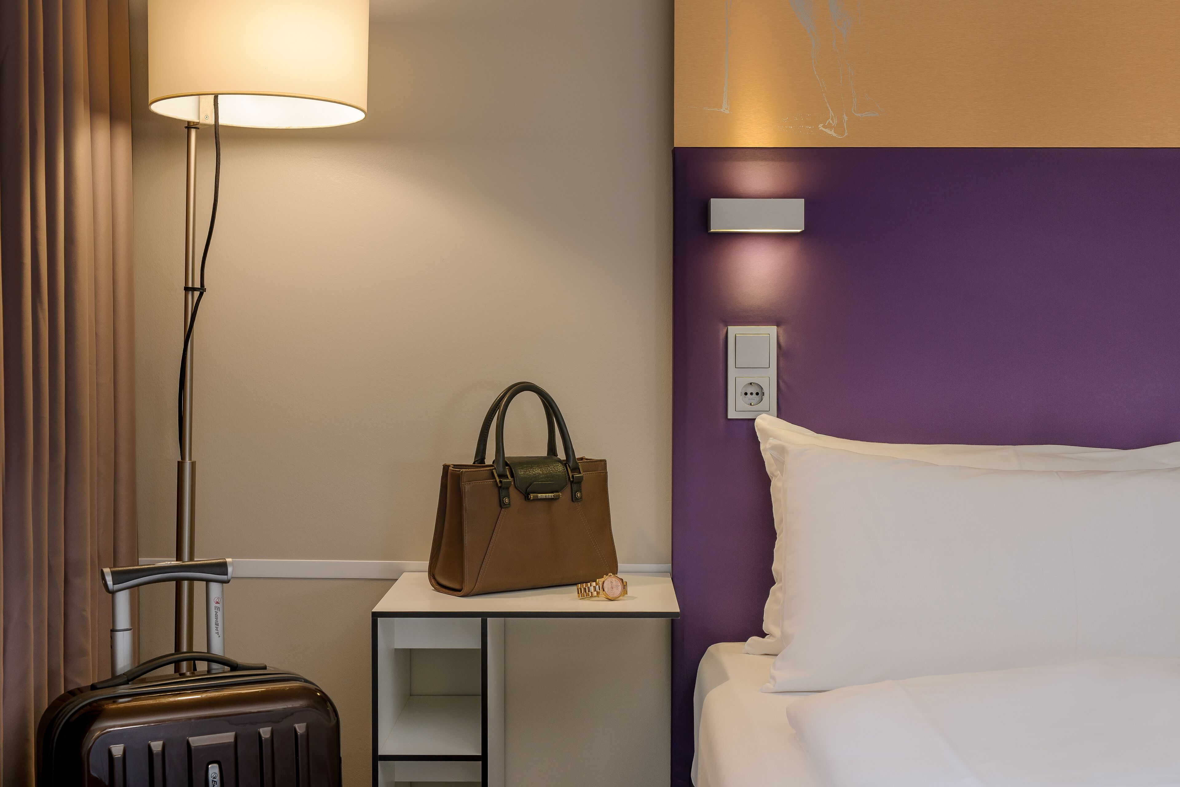 Una habitación de hotel con una cama, una mesita de noche, una lámpara, una maleta y un bolso.