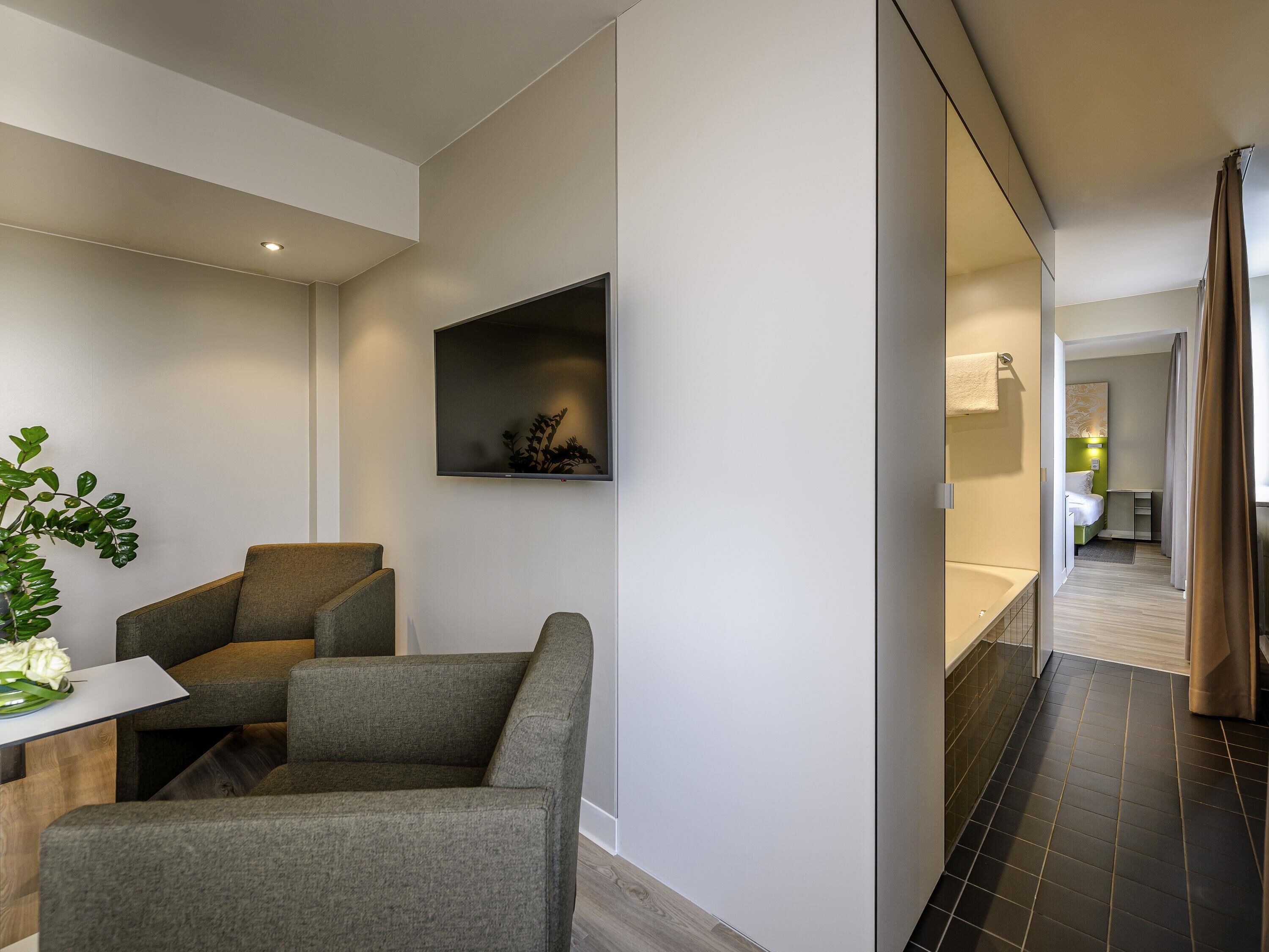A modern hotel room with a seating area, a flat-screen TV, a small shelf with decorative items, and a view into the bathroom.