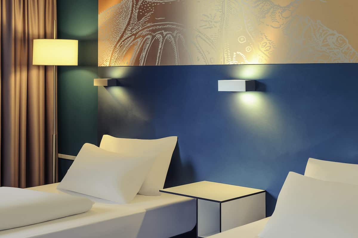 Habitación de hotel con dos camas, una mesita de noche, una lámpara y una pared con diseño decorativo.