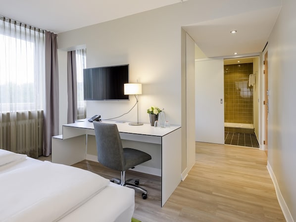 A hotel room with a bed, a desk with a chair, a TV, a lamp, and a shower area. - Congress Hotel Mercure Nürnberg an der Messe (Nuremberg)