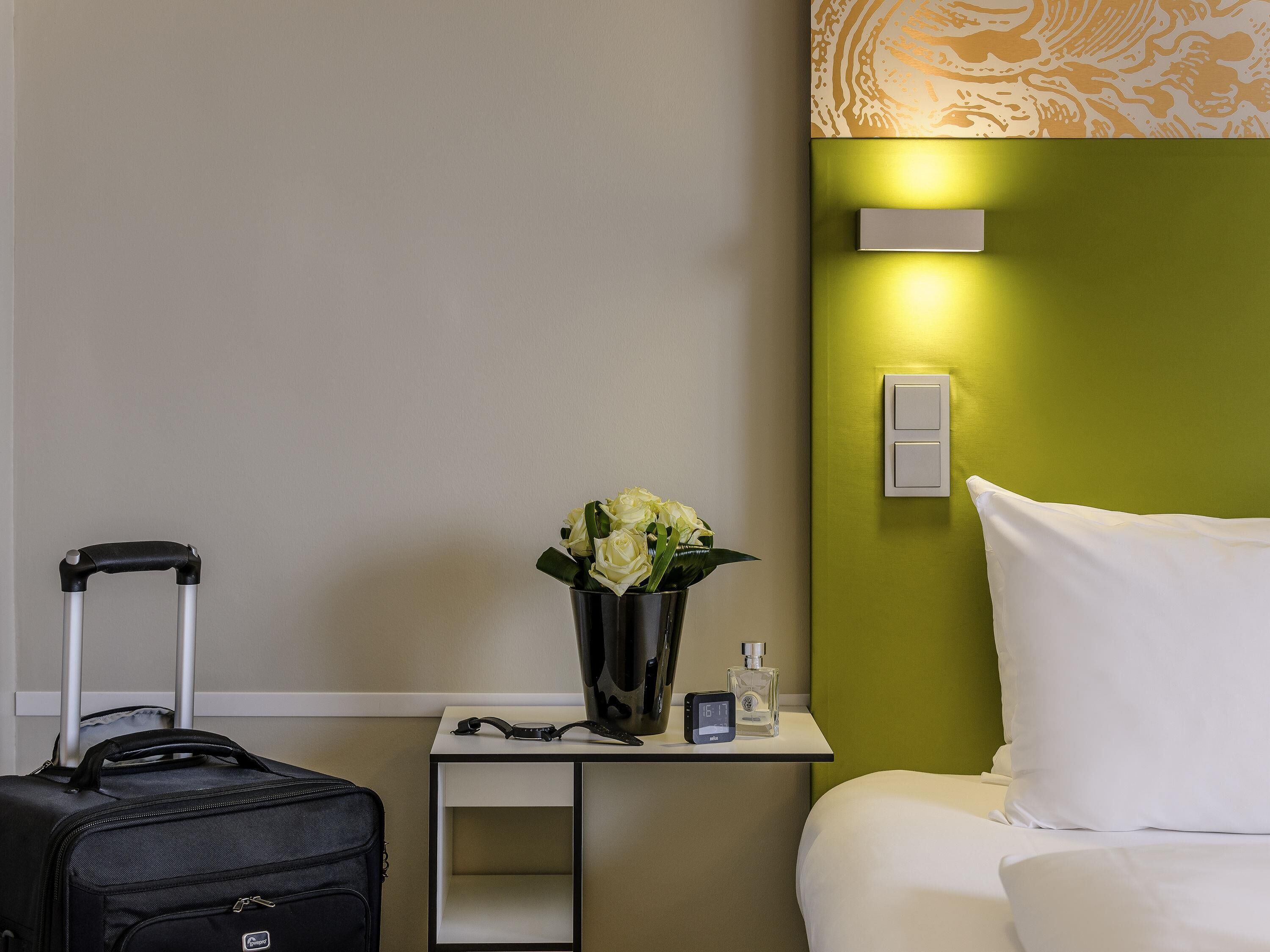 A hotel room with a bed, a nightstand with a vase of flowers, a suitcase, and a digital clock.