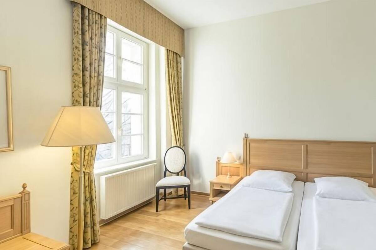 Habitación de hotel con cama, una silla, lámpara, mesita de noche y ventana con cortinas.