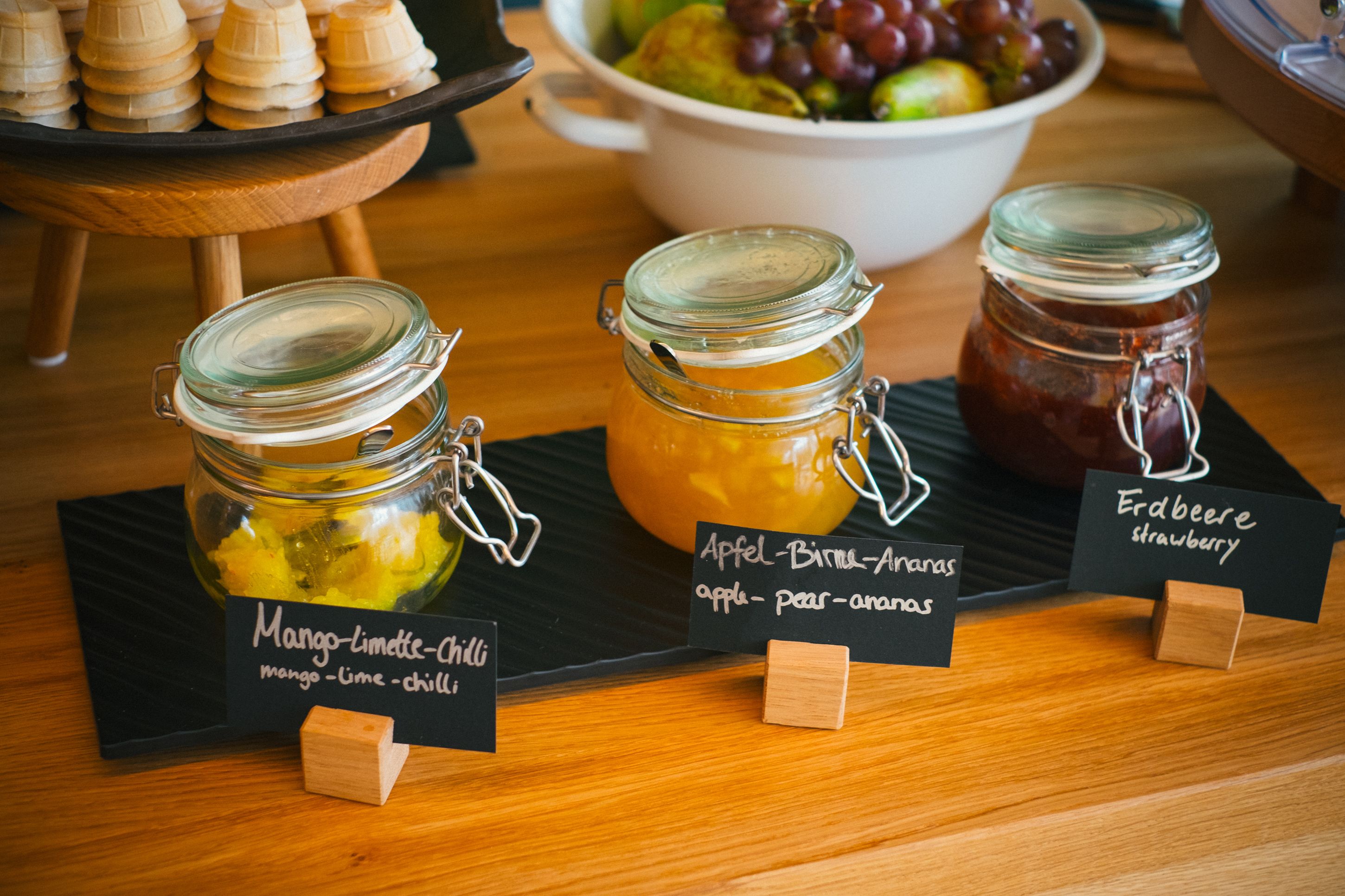 Tres botes de conservas etiquetados con mango-lima-chile, manzana-birne-anas y erdberee fresa sobre una mesa de madera.