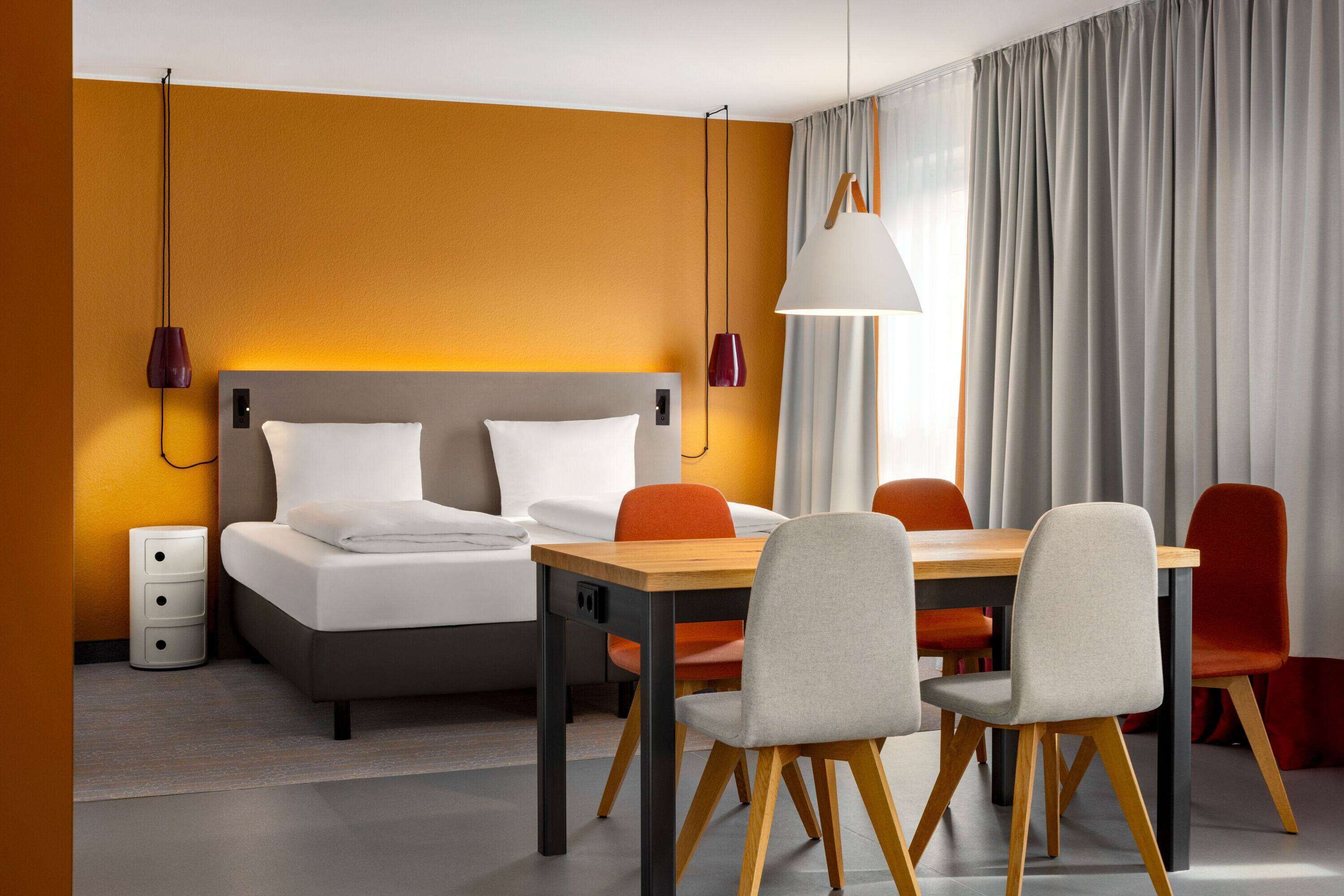 Moderni hotellihuone, jossa on sänky, puinen ruokapöytä ja neljä tuolia. Huoneessa on yöpöytä, jossa on laatikoita ja lamppu.