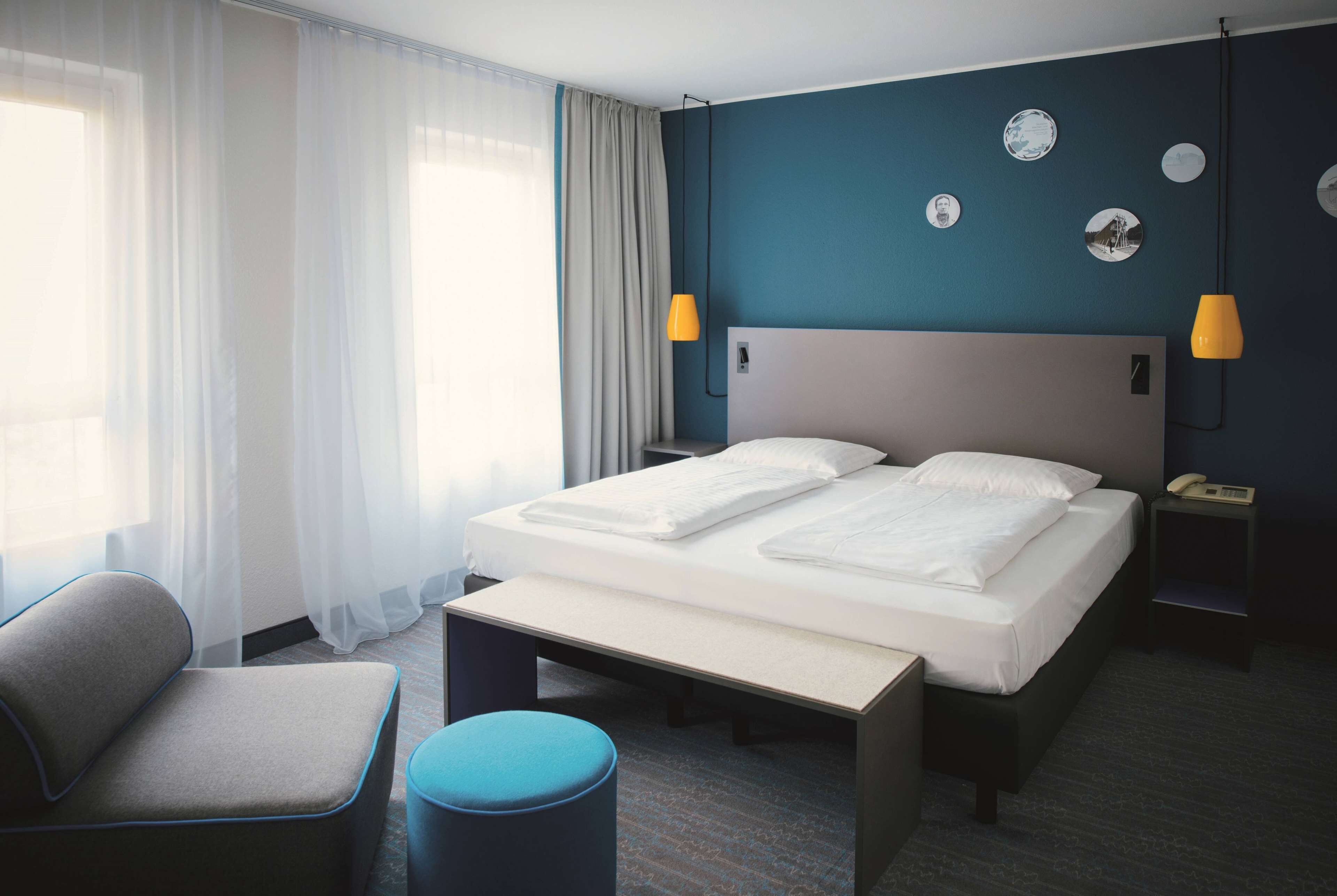 A hotel room with a bed, bedside tables, a chair, a bench, and a large window with curtains.