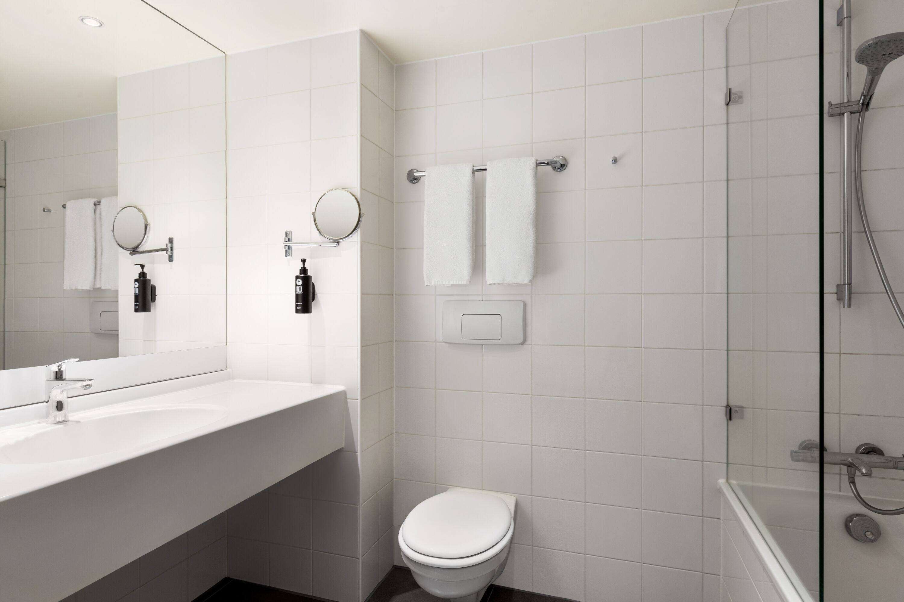 Un baño moderno con un lavabo blanco, un inodoro, ducha con mampara de vidrio y dos toallas blancas en un perchero.