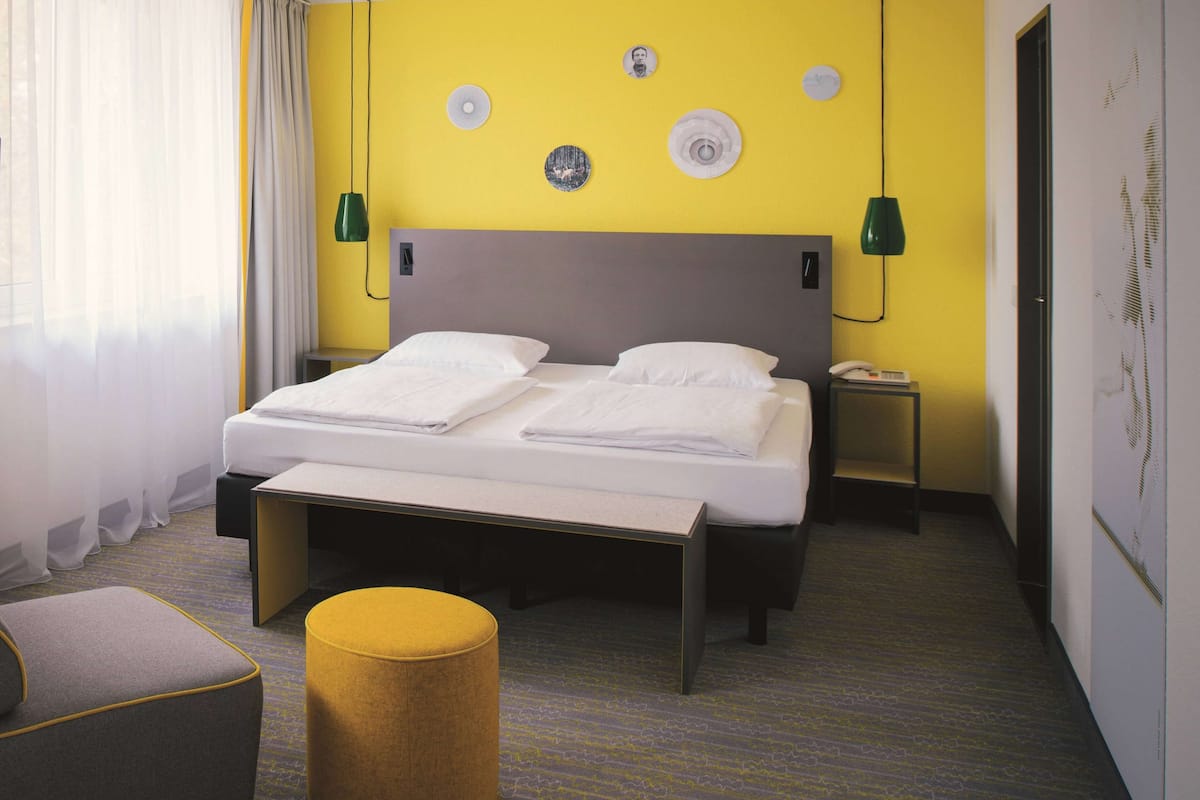 A hotel room with a bed, bedside table, lamp, and a small stool.