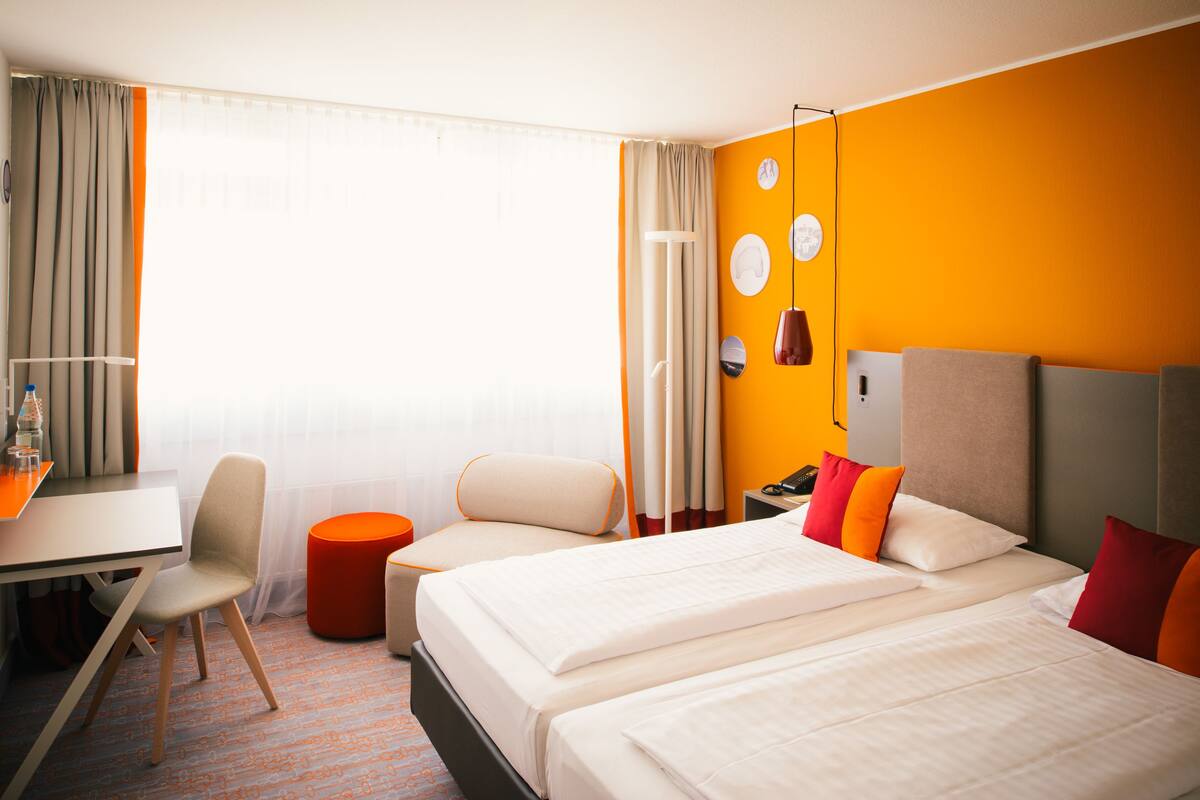 A hotel room with two beds, a desk, a chair, and a small orange ottoman.