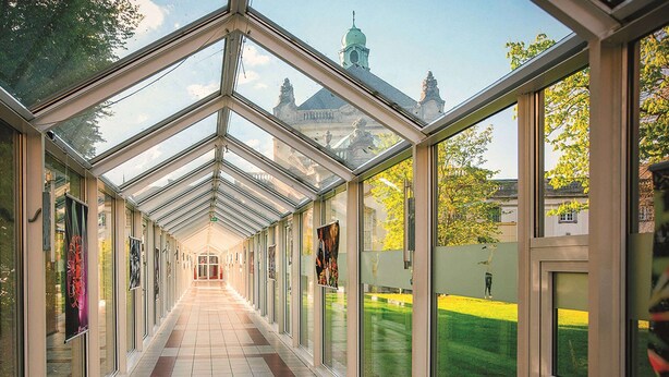 A covered corridor with glass walls, a tiled floor, and posters on the walls. - Vienna House Easy by Wyndham Bad Oeynhausen (Bad Oeynhausen)