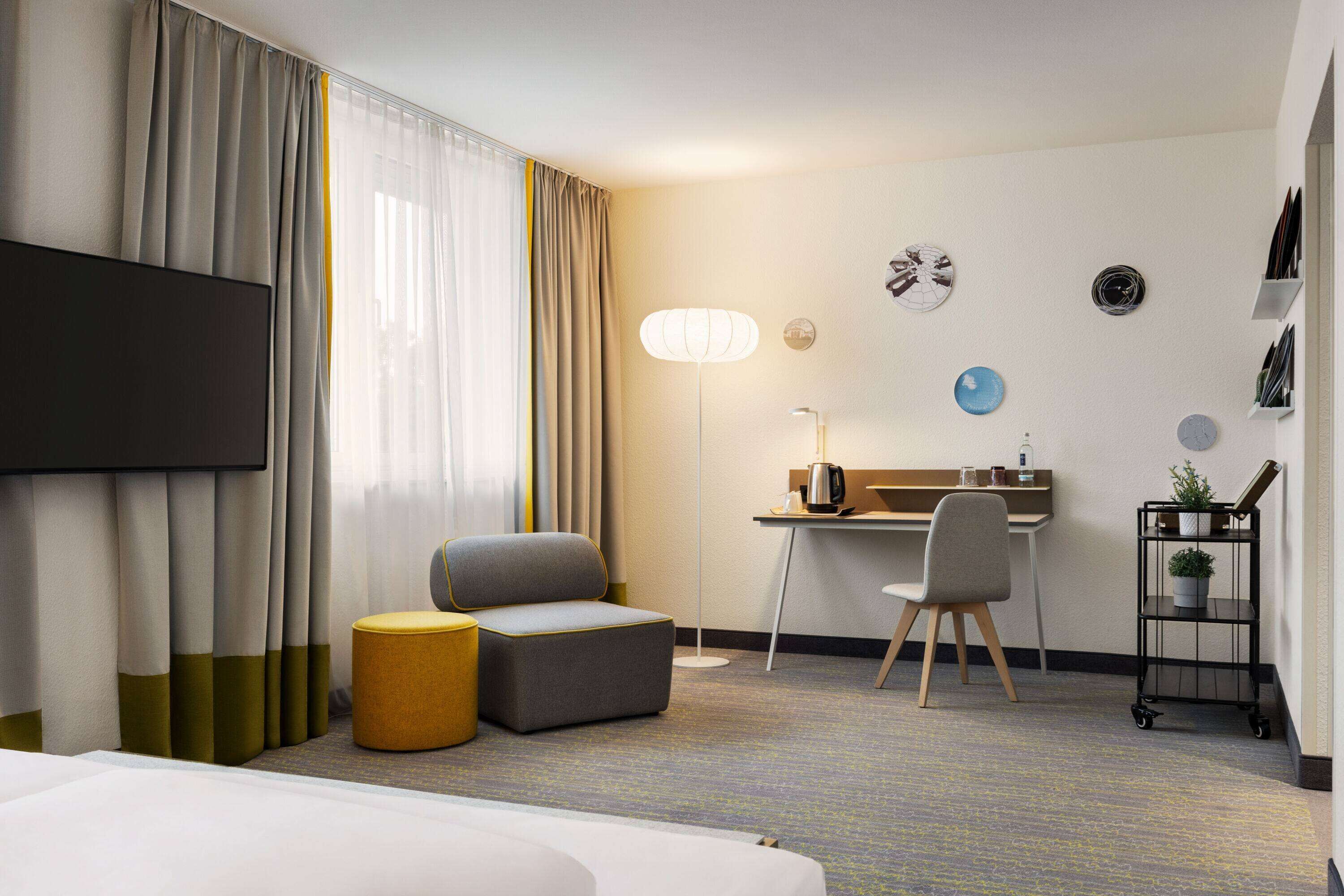 Een hotelkamer met een bed, een bureau, een stoel, een klein voetenbankje, een televisie, een lamp, een klok en een plankje met een plant.