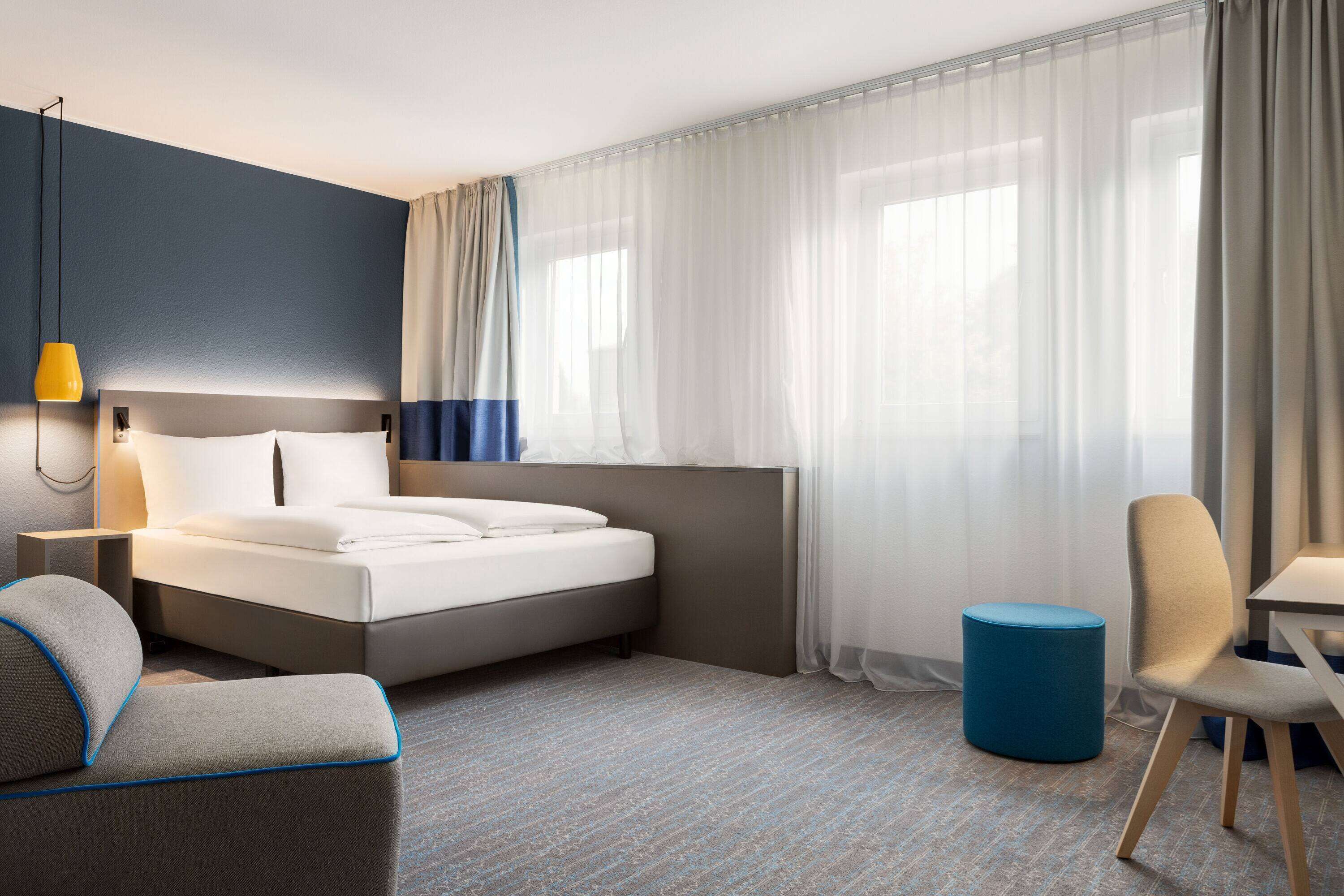 A modern hotel room with a bed, a desk, a chair, and a small ottoman.
