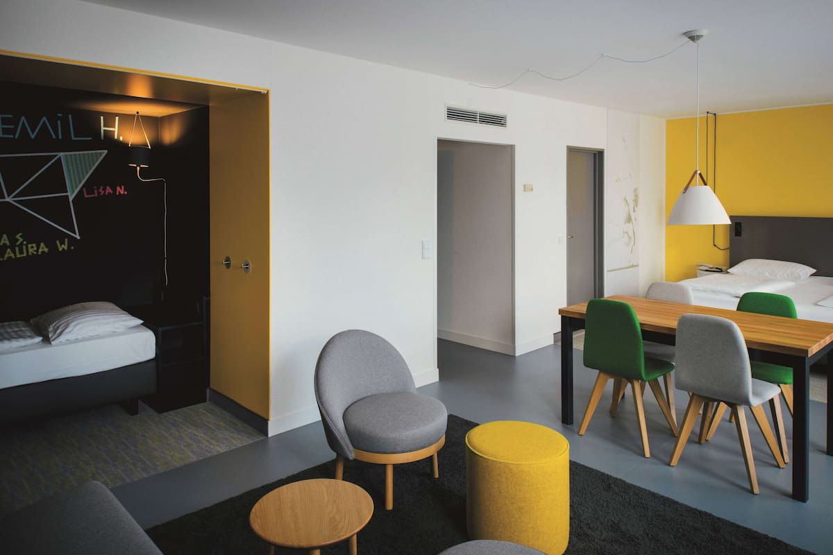 A modern hotel room with a bed, a desk, and chairs. There is a yellow lamp, a black wall with text, and a white door.