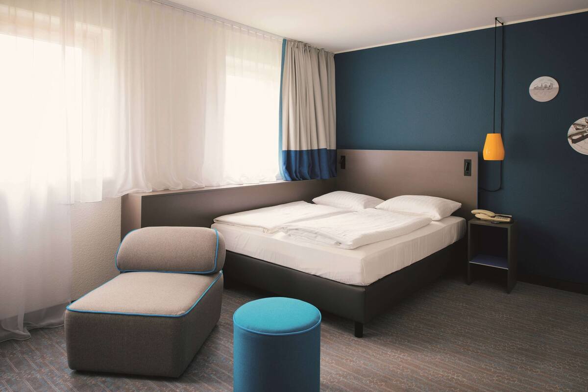 A modern hotel room with a bed, a chair, a small table with a lamp, and a window with curtains.