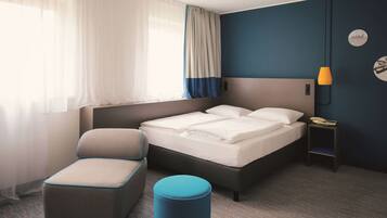 Ein modernes Hotelzimmer mit Bett, Stuhl, kleinem Tisch mit Lampe und einem Fenster mit Vorhängen.