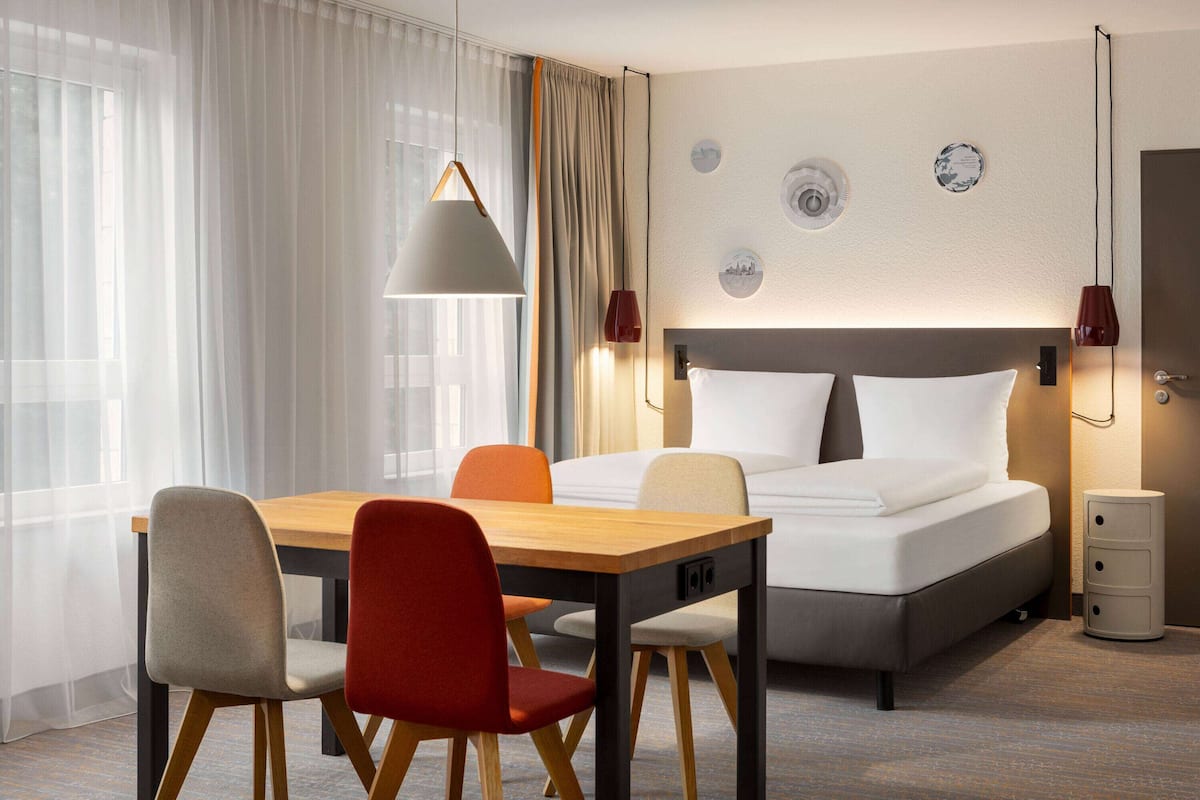 A hotel room with a bed, a wooden table, and two chairs. There is a bedside table with a lamp and a small drawer unit.