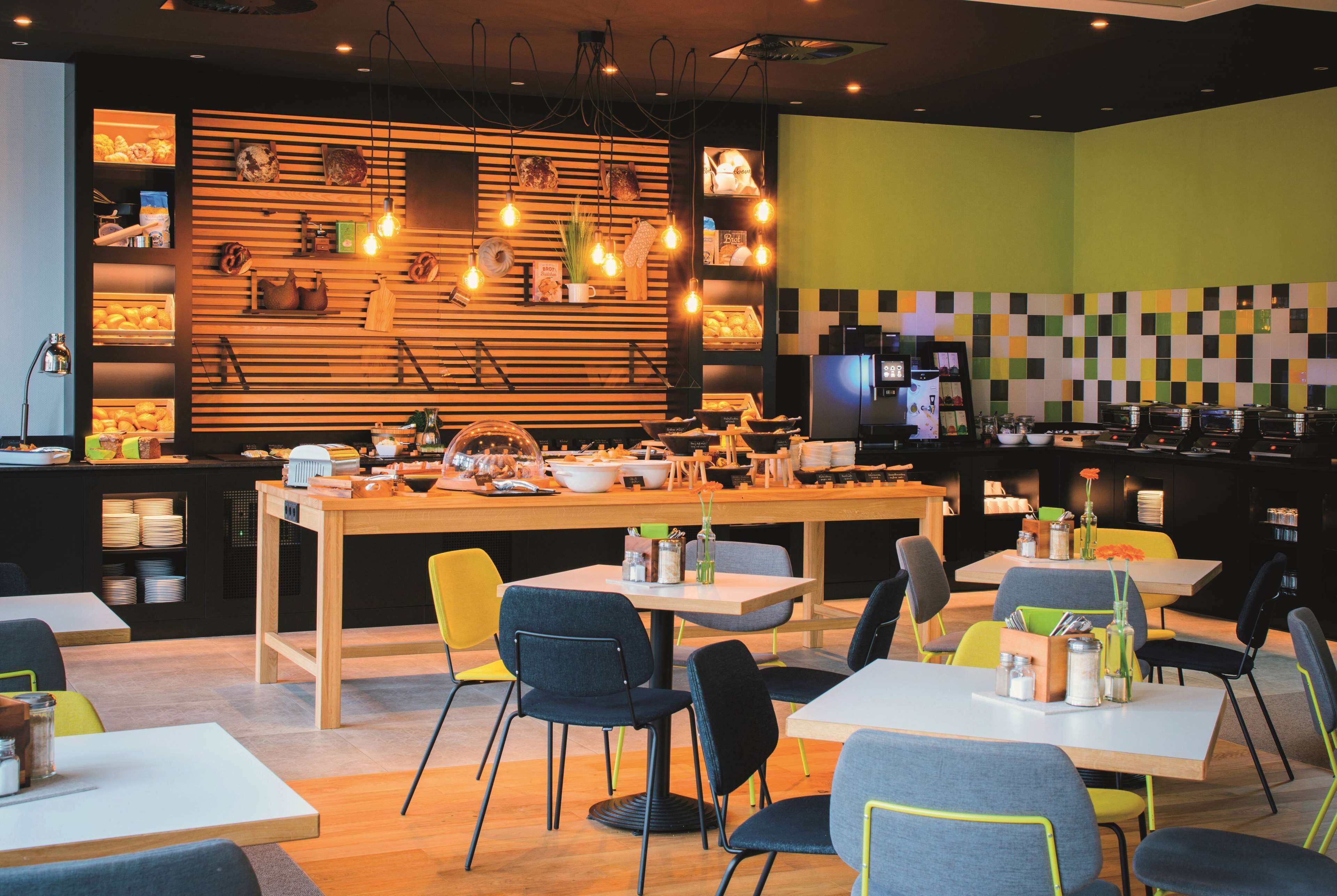 A modern restaurant with a central wooden table, surrounded by chairs, and a kitchen area with appliances and shelves in the background.