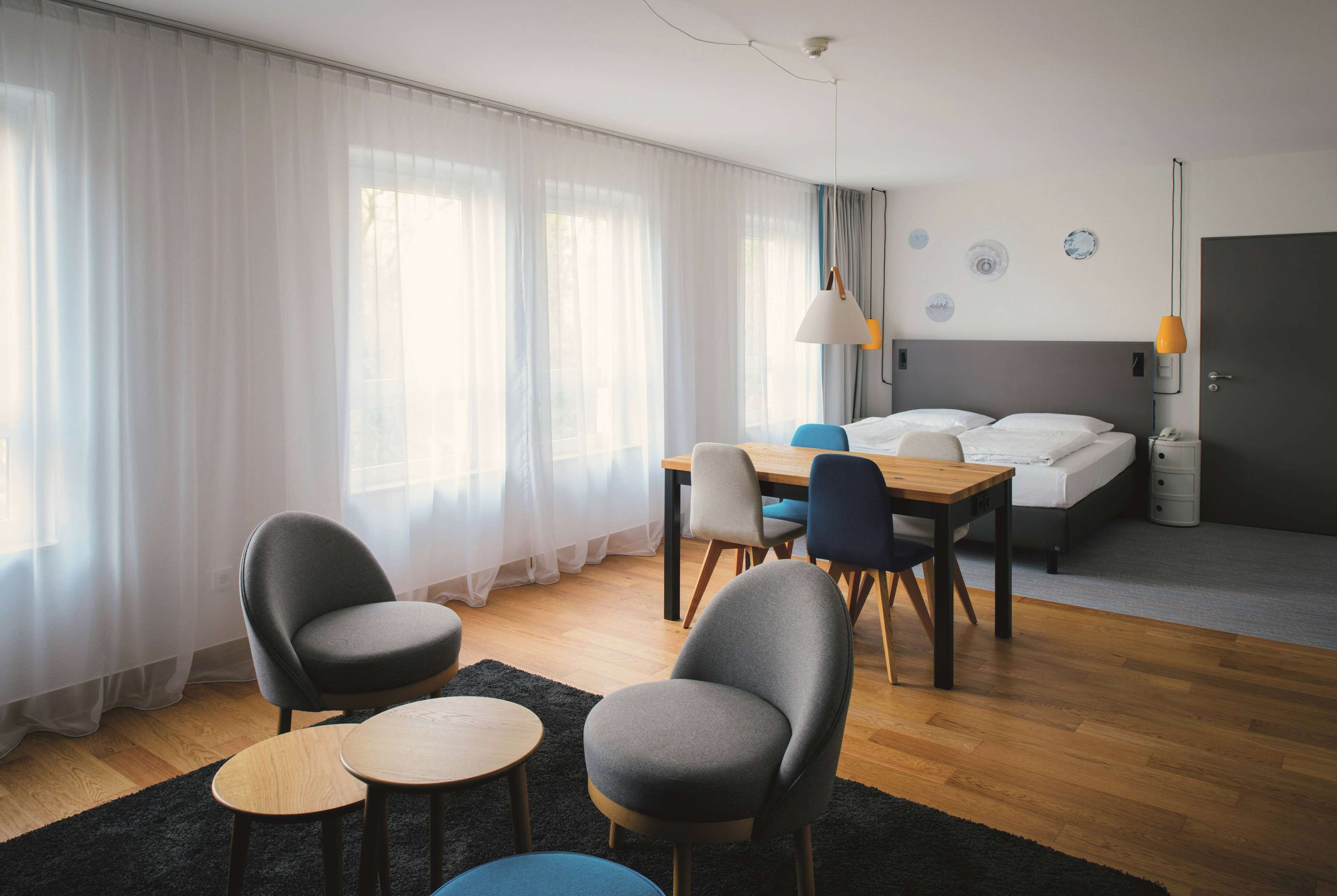 A modern hotel room with a wooden floor, a bed with white linens, a desk with chairs, and a large window with sheer curtains.