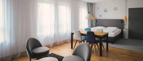 A modern hotel room with a wooden floor, a bed with white linens, a desk with chairs, and a large window with sheer curtains.