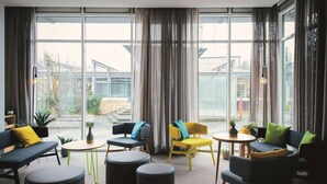 A modern lounge area with a sofa, chairs, and ottomans arranged around a central table. Large windows provide a view of the outside. - Vienna House Easy by Wyndham Bad Oeynhausen (Bad Oeynhausen)
