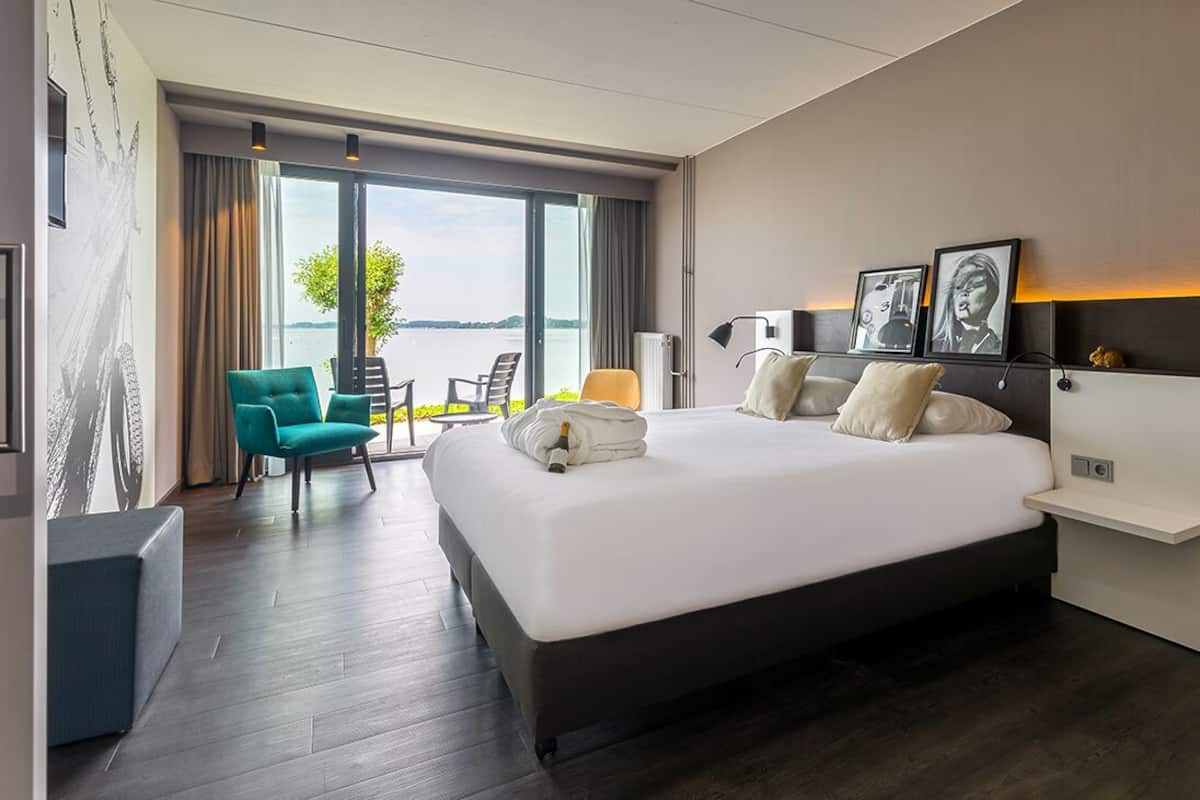 Une chambre d’hôtel moderne dotée d’un grand lit, d’un coin salon avec un fauteuil, d’un bureau et offrant une vue sur l’extérieur.