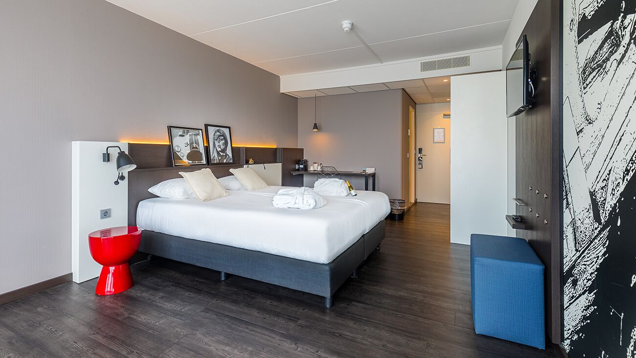 A modern hotel room with a large bed, a red toilet, a small table, and a blue bench.