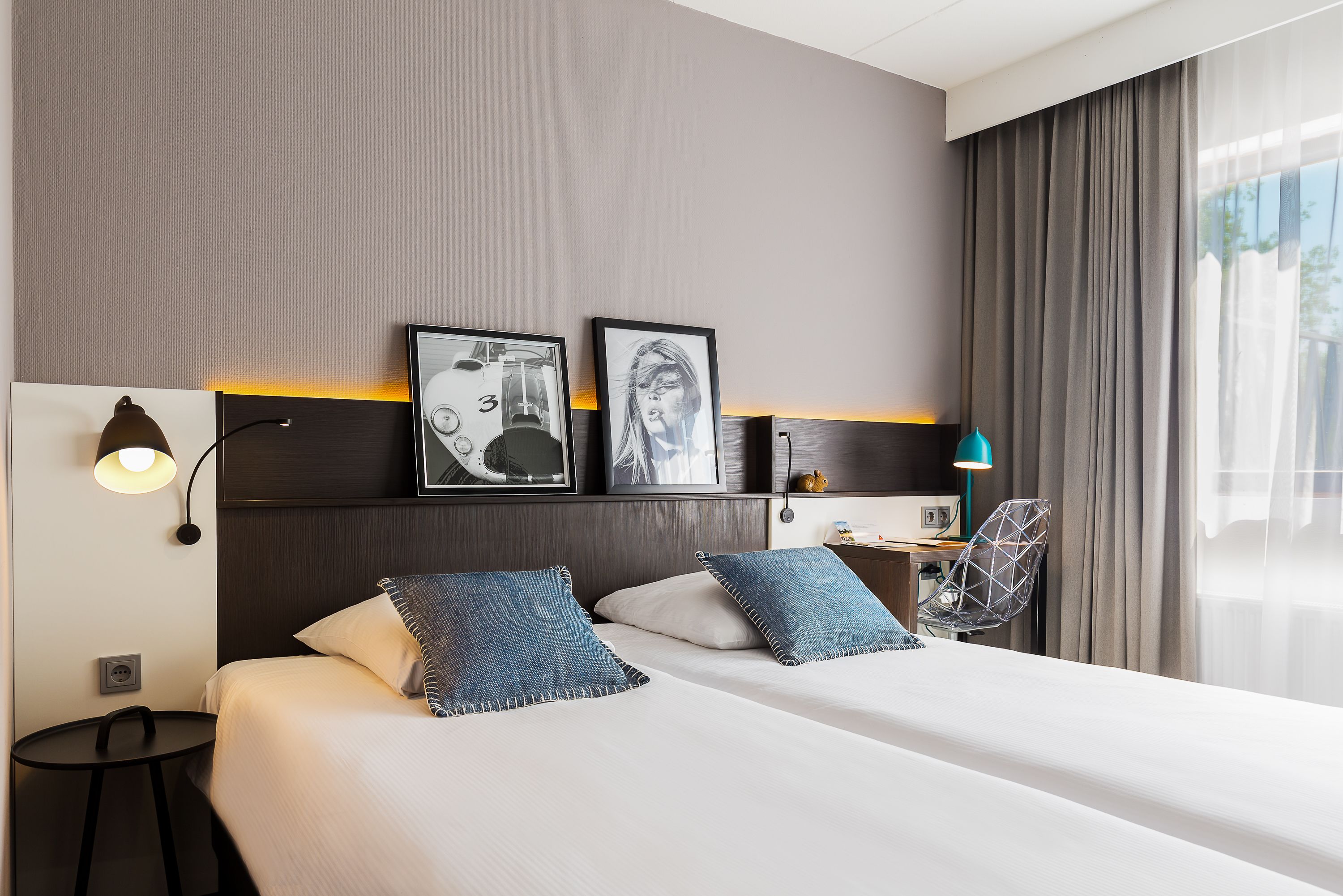 A hotel room with a large bed, two pillows, a nightstand, a lamp, and a wall-mounted shelf with framed pictures.