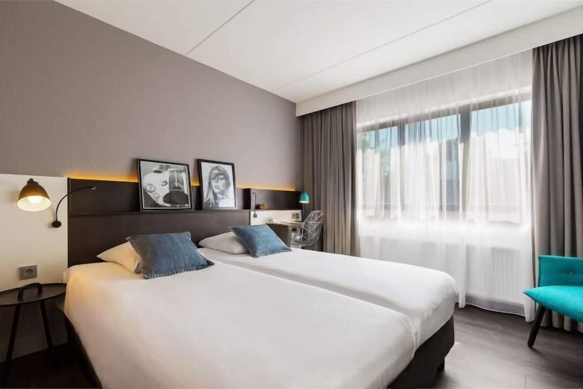 Une chambre d’hôtel avec deux lits, un bureau, une chaise et une grande fenêtre avec des rideaux.