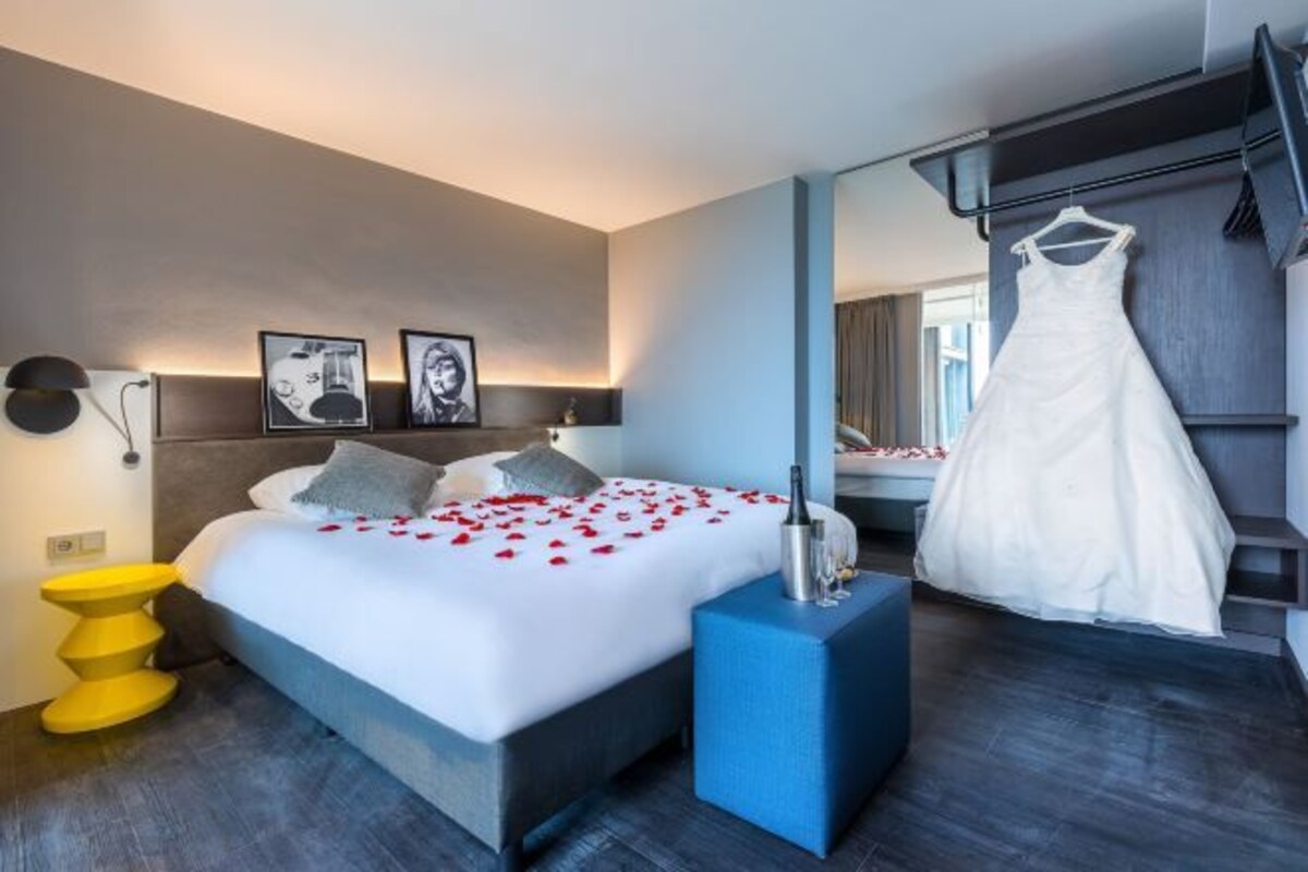 Une chambre d’hôtel moderne avec un grand lit, un élément décoratif bleu, une robe suspendue et une lampe fixée au mur.