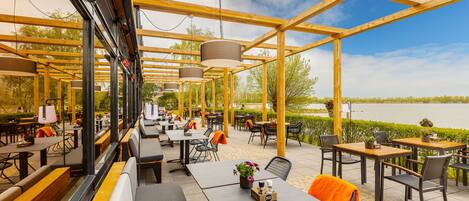 Ein Restaurant mit Sitzbereich im Freien, Holztische und -stühle, Blick auf einen See und ein überdachter Bereich mit hängenden Lichtern.