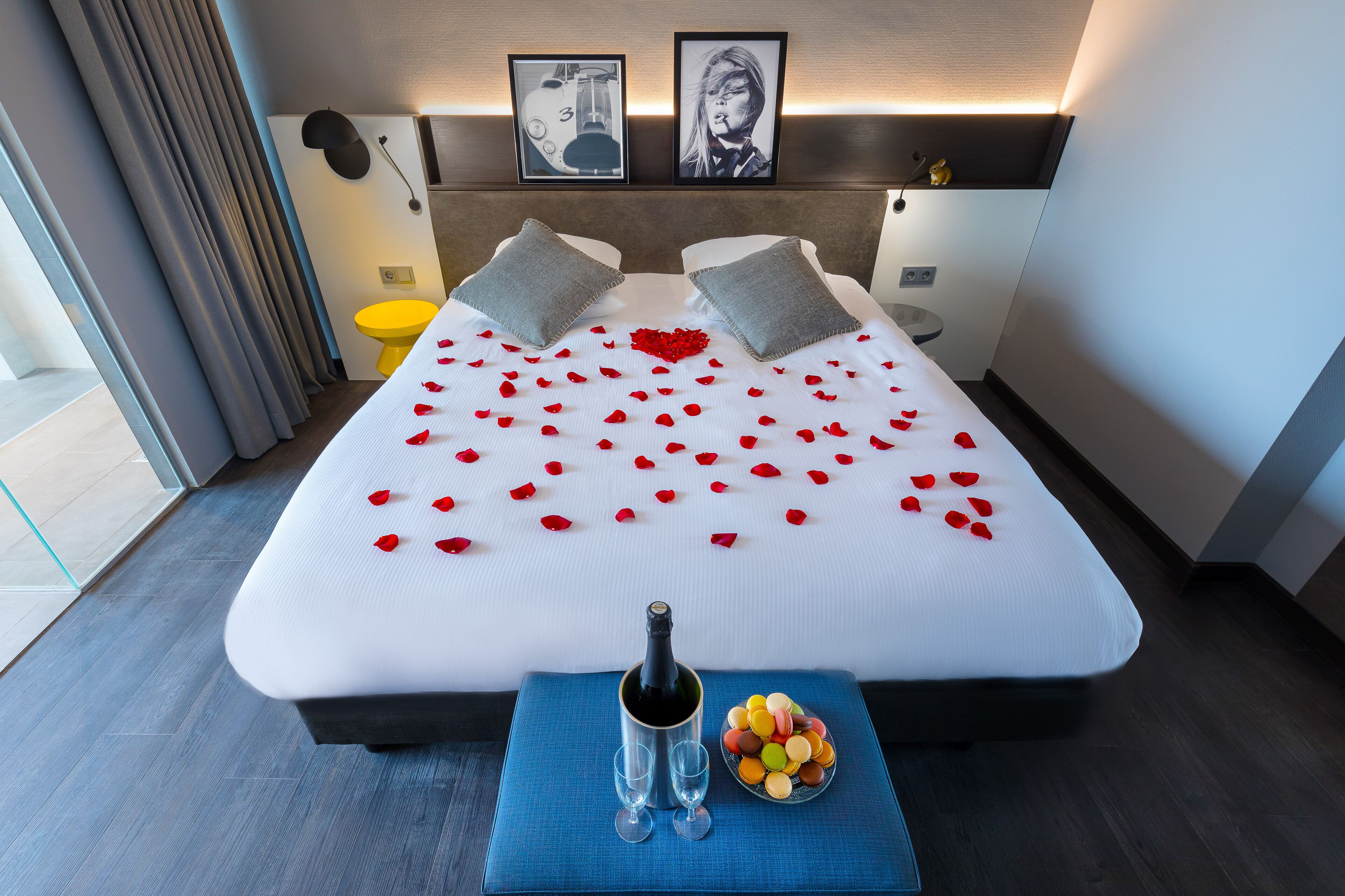 Een hotelkamer met een bed versierd met rozenblaadjes, een fles champagne en een schaaltje macarons.