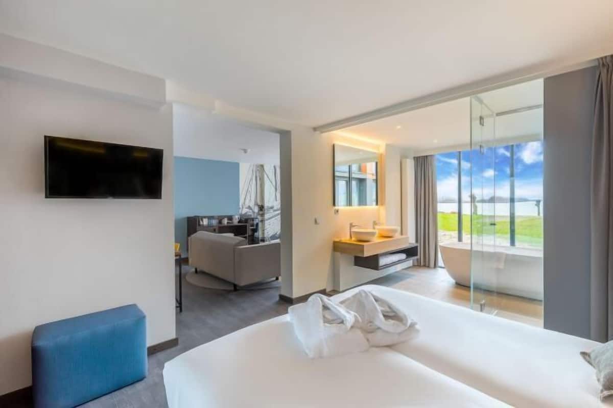 Une chambre d’hôtel moderne équipée d’un lit, d’une télévision, d’un bureau avec une chaise, d’une salle de bain avec un lavabo et d’une grande fenêtre donnant sur un paysage agréable.