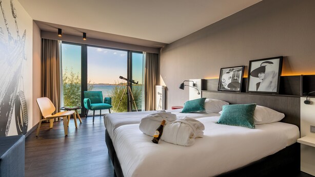 Une chambre d’hôtel avec deux lits, une grande fenêtre donnant sur un beau paysage, un fauteuil vert et une table sur laquelle se trouve une bouteille de champagne.
