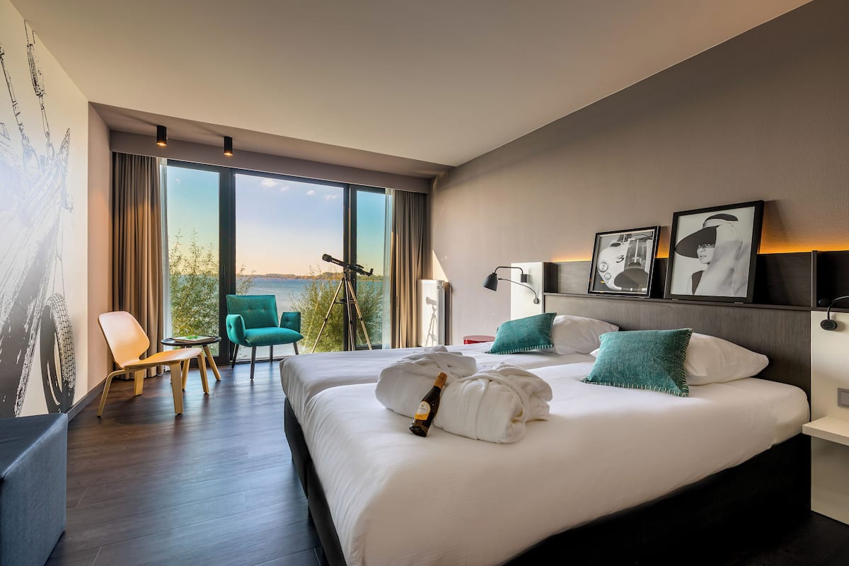 Une chambre d’hôtel avec deux lits, une grande fenêtre donnant sur un beau paysage, un fauteuil vert et une table sur laquelle se trouve une bouteille de champagne.