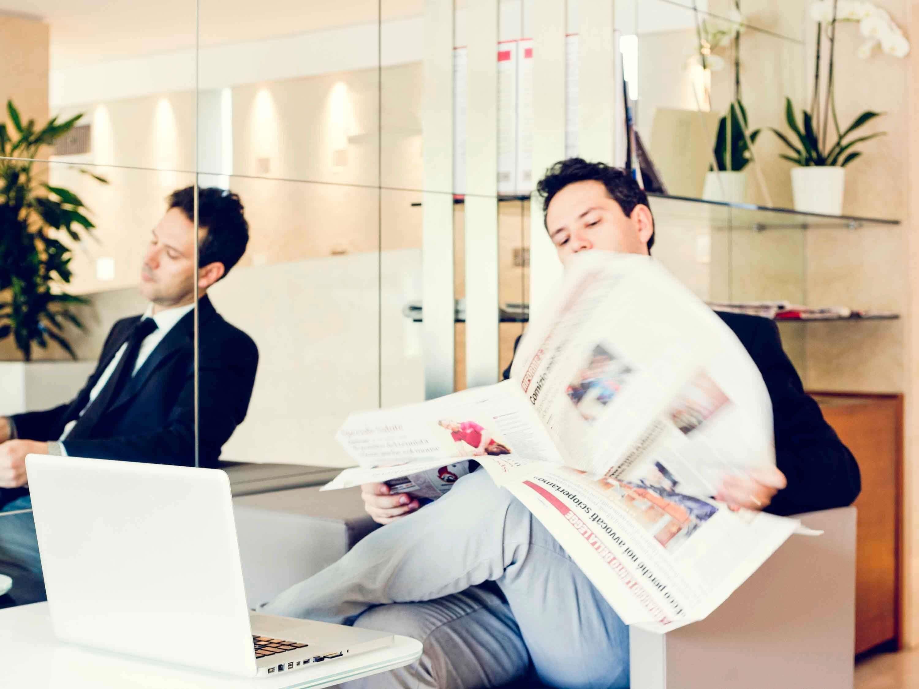 Un ambiente d'ufficio moderno con pareti divisorie in vetro, un portatile su un tavolo e una persona che legge un giornale.