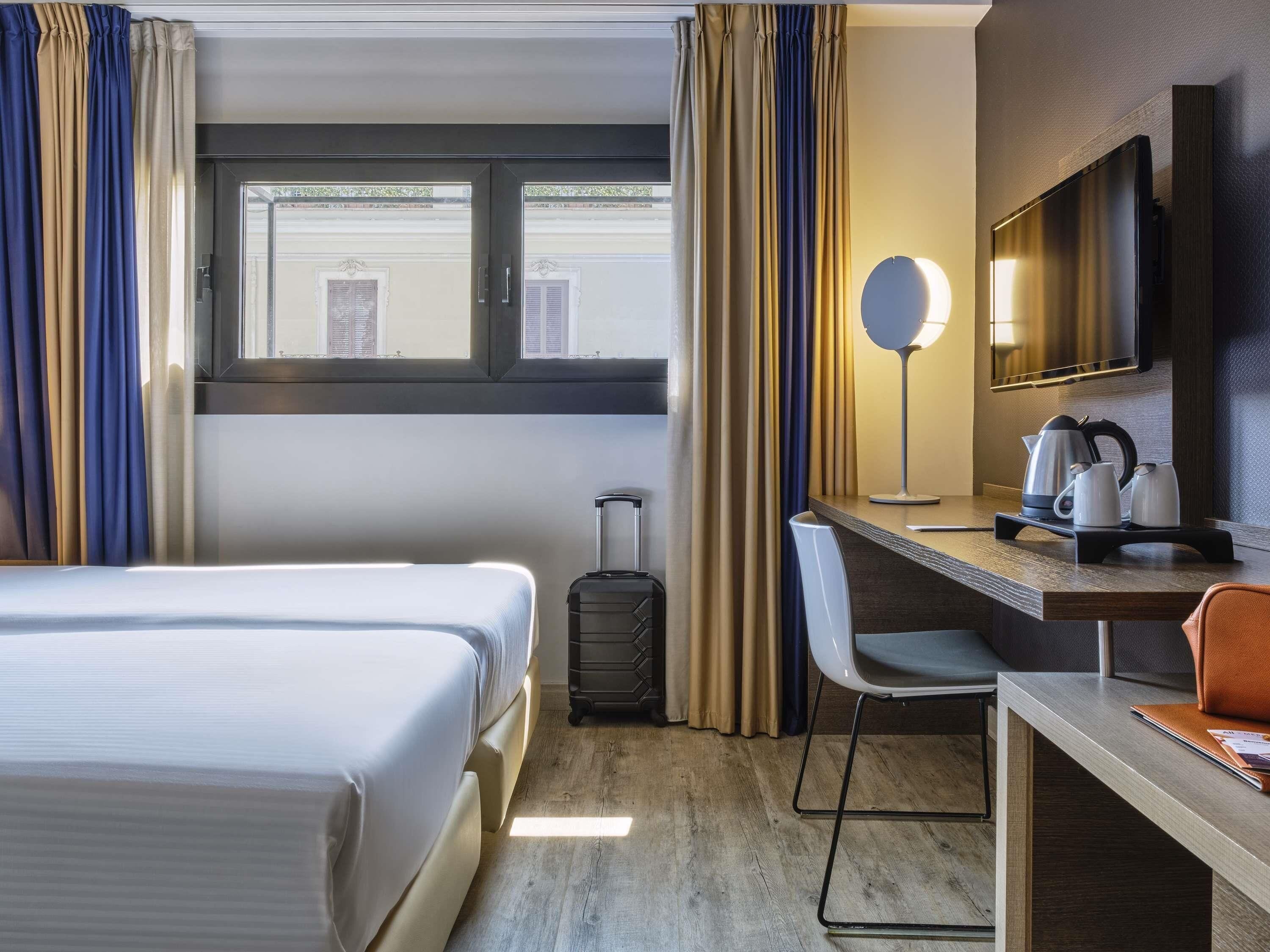 A hotel room with a bed, a desk with a chair, a suitcase, a flat-screen TV, and a window with curtains.