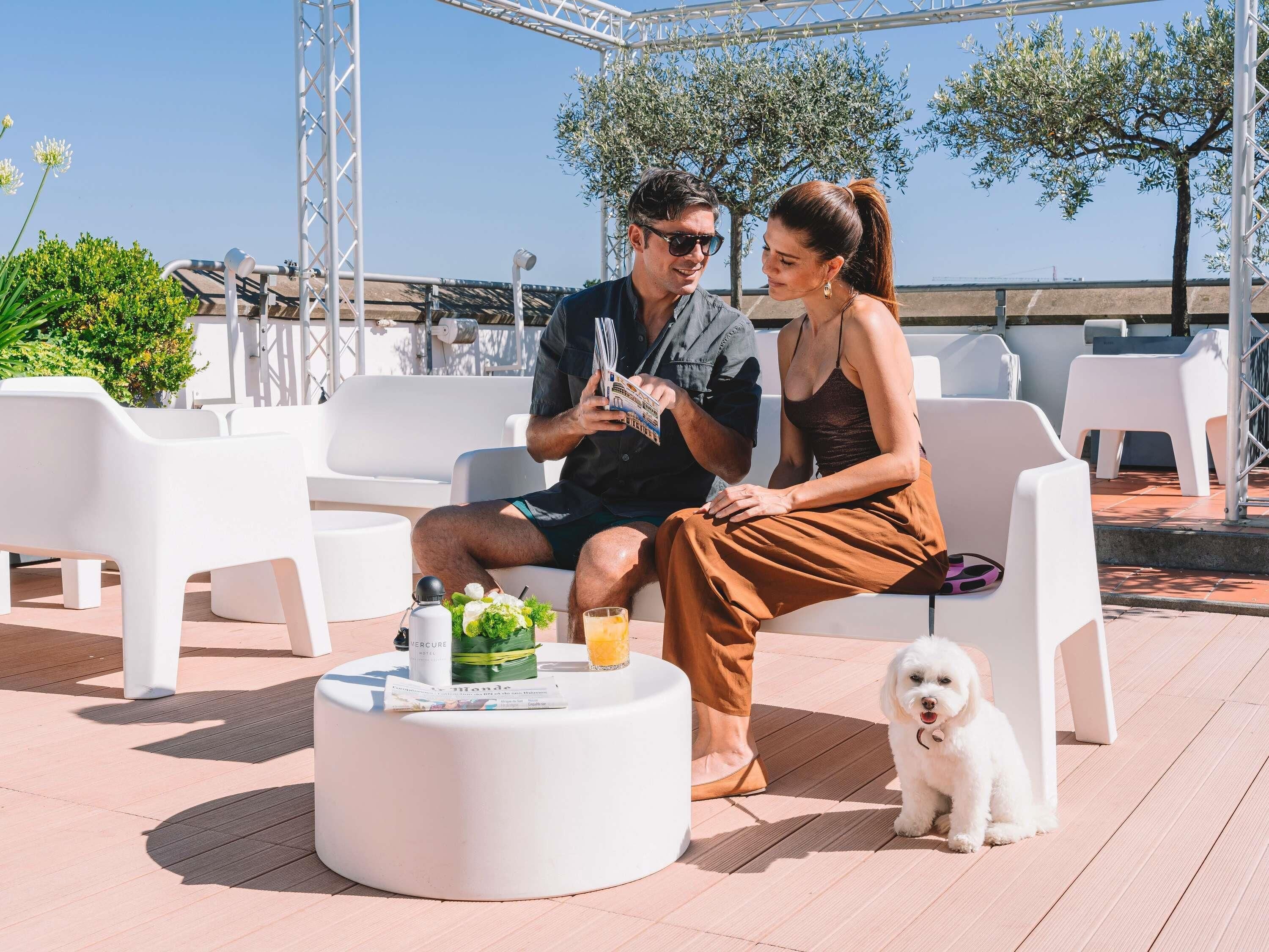 Two people sitting on white outdoor chairs, a small round table with drinks and a bottle, and a white dog.