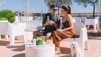 Two people sitting on white outdoor chairs, a small round table with drinks and a bottle, and a white dog.