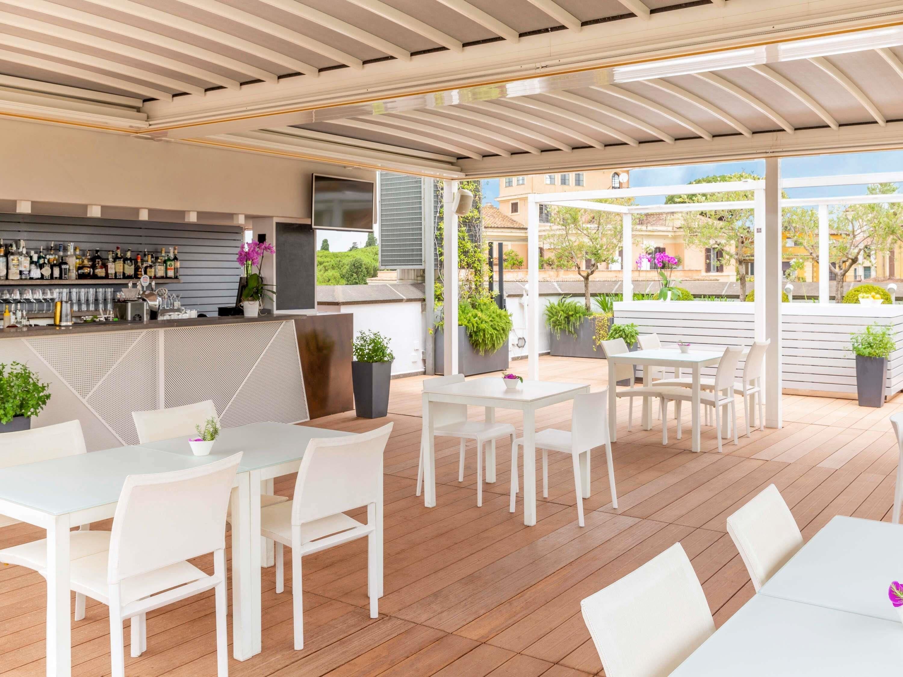 An outdoor patio area with white tables and chairs, a bar with various bottles, and potted plants.