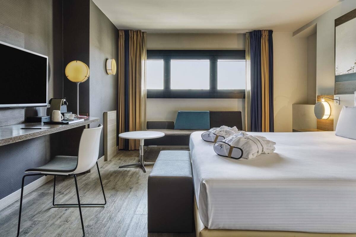 Une chambre d’hôtel comprenant un lit, un bureau avec une chaise, une petite table, un téléviseur et une fenêtre avec des rideaux.