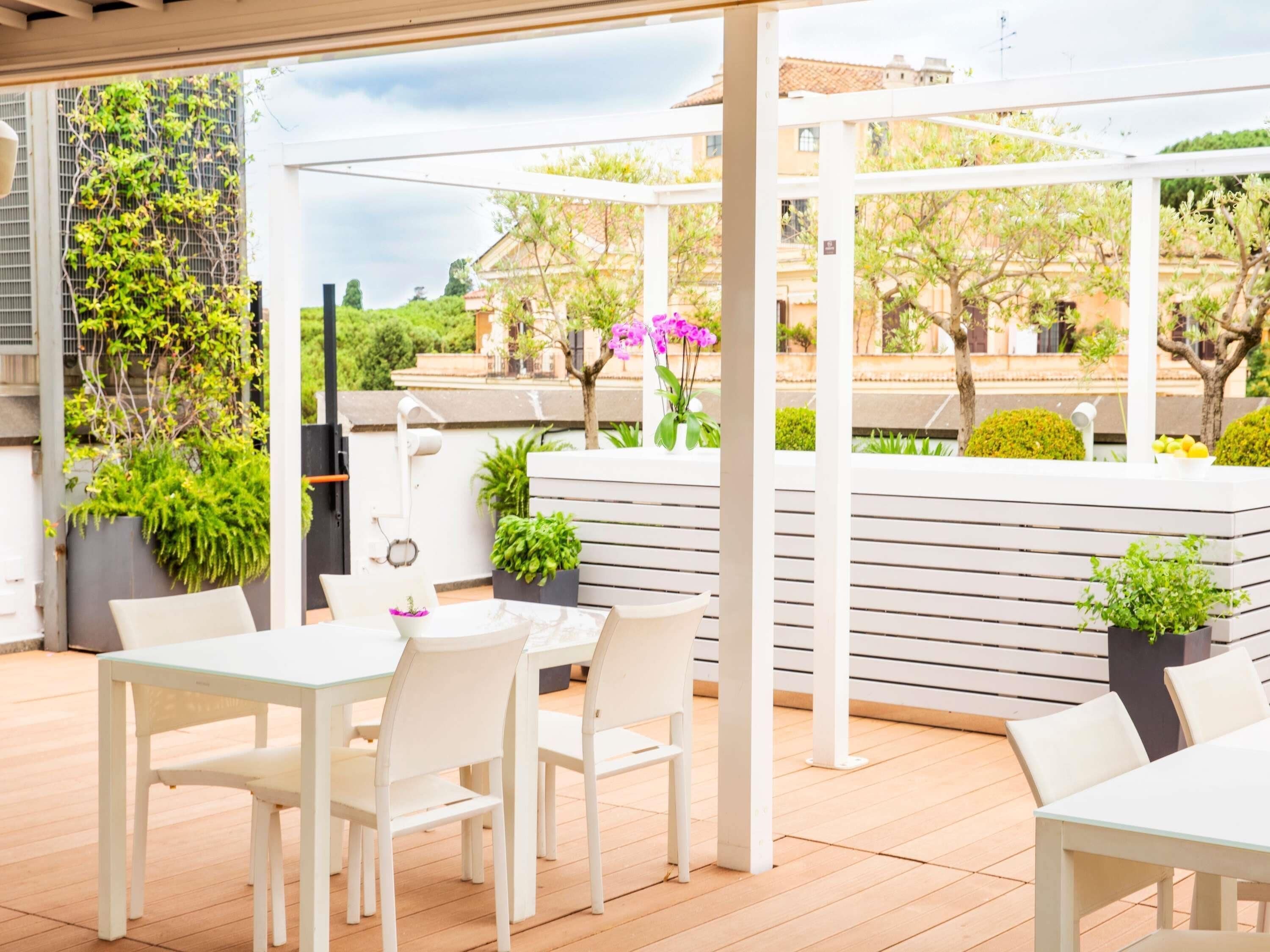 An outdoor patio area with white furniture, including tables and chairs, surrounded by potted plants and a pergola structure.