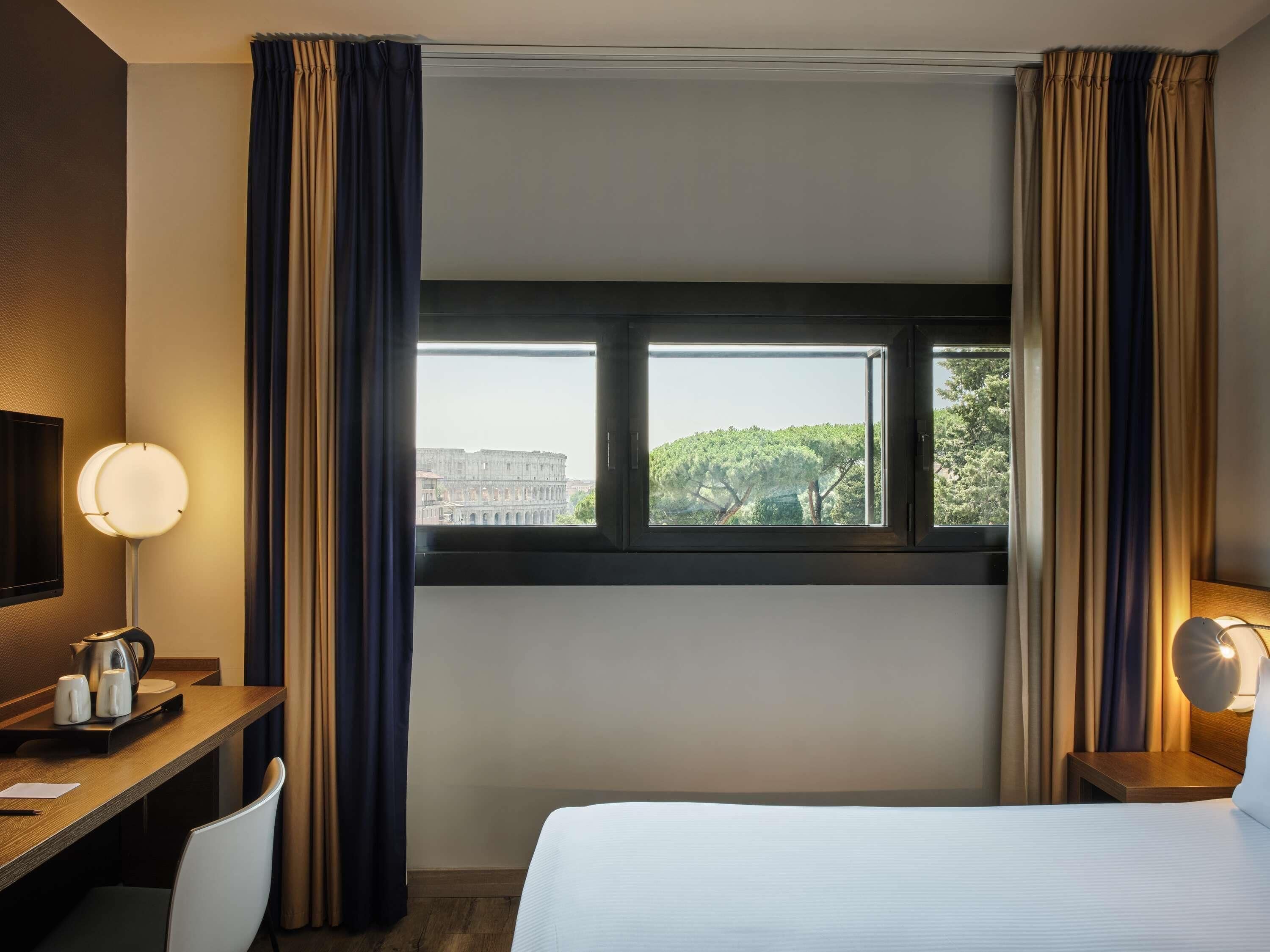 A hotel room with a bed, a desk, a chair, a nightstand, a lamp, and a window with curtains.