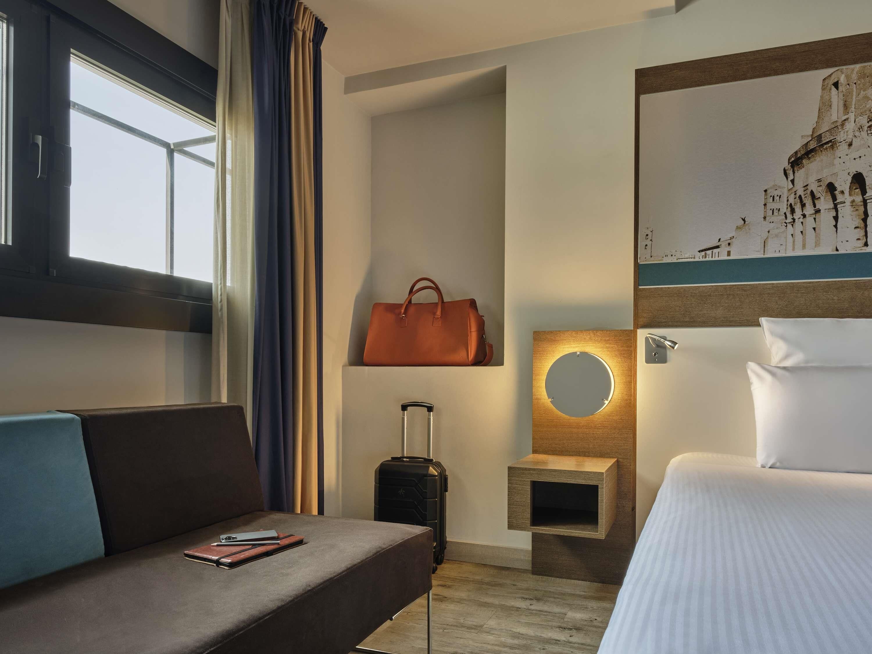Ett hotellrum med en säng, en soffa, ett litet bord, en spegel som hänger på väggen, en resväska och en målning av en historisk byggnad.
