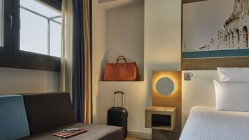 A hotel room with a bed, a sofa, a small table, a wall-mounted mirror, a suitcase, and a painting of a historical building.