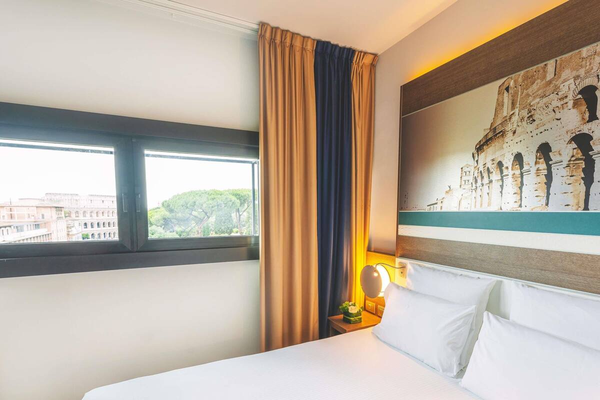 Une chambre d’hôtel avec un lit, une fenêtre avec des rideaux, une table de chevet avec une lampe et un mur orné d’une fresque.