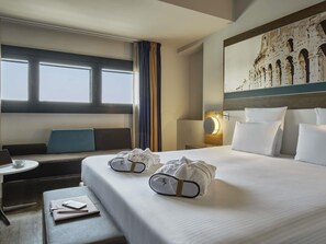 A hotel room with a large bed, a desk with a laptop, a chair, and a large window.